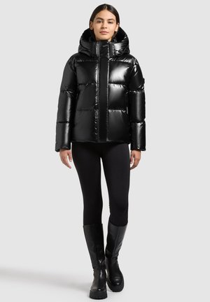 Joven mujer que lleva una chaqueta acolchada negra brillante, mallas negras y botas negras hasta la rodilla, de pie contra un fondo liso.