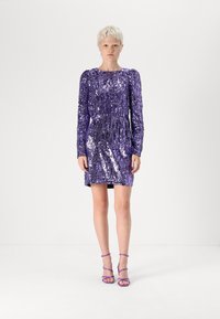 Vestido roxo de lantejoulas com mangas compridas, corpete justo e textura pregueada. Combinado com saltos roxos com tiras. Fundo de estúdio.