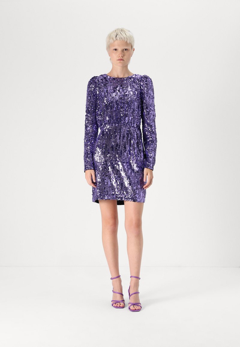 Vestido roxo de lantejoulas com mangas compridas, corpete justo e textura pregueada. Combinado com saltos roxos com tiras. Fundo de estúdio.