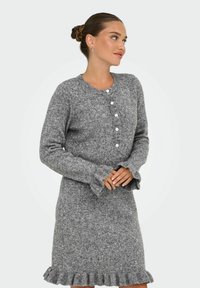 Femme dans une robe en tricot grise avec des boutons blancs, des poignets froncés et un ourlet froncé, se tenant les mains jointes et regardant sur le côté.