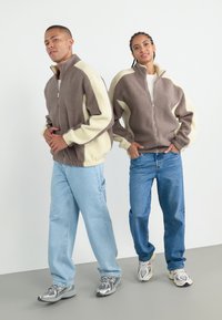 Twee modellen dragen ritsjackets van zachte, gestructureerde fleece. De jassen zijn bruin met crèmekleurige accenten. Beide dragen casual jeans en sneakers.