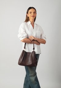 Borsa a mano in pelle marrone con una forma strutturata, doppi manici e dettagli di cucitura sottile. Indossata con una camicia bianca e jeans blu.