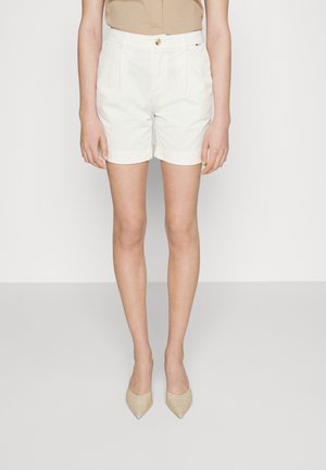 Mujer con pantalones cortos blancos a medida de talle alto con botón y cremallera, combinados con zapatos beige de punta afilada y una blusa marrón claro.