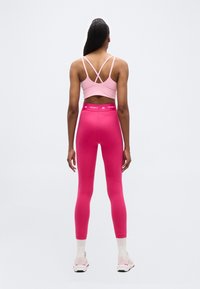 Γυναίκα που στέκεται με την πλάτη γυρισμένη, φοράει ροζ αθλητικό σουτιέν, φωτεινά ροζ ψηλόμεσα κολάν, λευκές κάλτσες και ροζ-λευκά αθλητικά παπούτσια πάνω σε λευκό φόντο.