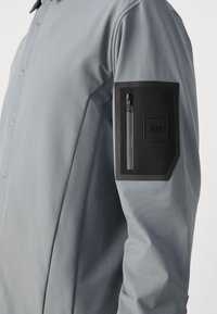 Veste grise à boutons avec un tissu texturé, des manches longues et une poche zippée noire sur le bras gauche avec un patch de logo.