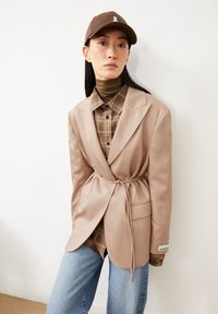 Blazer beige con un design a cintura, indossato sopra una camicia a quadri marrone e un dolcevita verde. Il modello indossa un berretto marrone con un logo bianco.