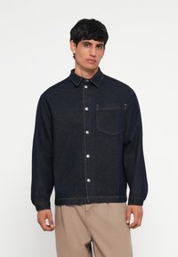 Temno modra denim jakna z gumbom spredaj, koničastim ovratnikom, enim žepom na prsih in manšetami. Ima srebrne gumbne okrase.