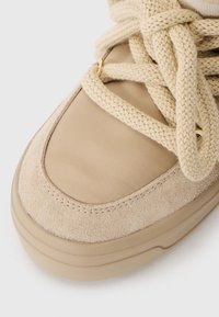 Beige sneaker featuring a mix of suede and textured fabric, round toe, thick laces, and gold eyelets for a secure fit.