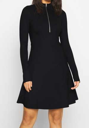 Kobieta ubrana w czarną prążkowaną sukienkę z długim rękawem, krótką spódnicą i zamkiem błyskawicznym z przodu od szyi do połowy klatki piersiowej.