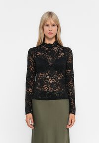 Woman with straight blonde hair wears a black floral lace long-sleeve top over a black bra and an olive-green skirt against a plain background.