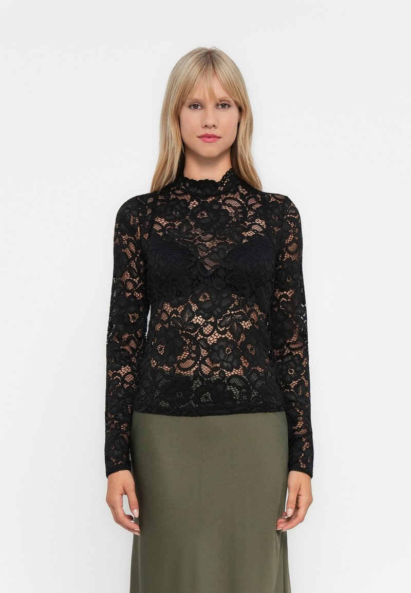 Woman with straight blonde hair wears a black floral lace long-sleeve top over a black bra and an olive-green skirt against a plain background.