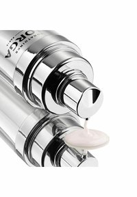 Silver cylindrical pump bottle with a glossy finish, dispensing a creamy, light pink product onto a smooth, white surface.
