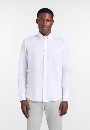 Homme noir debout, face à l’avant, portant une chemise blanche à manches longues boutonnée et un pantalon gris clair sur un fond uni.