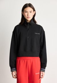 Black cropped sweatshirt with zip collar, featuring Calvin Klein logo in white on the chest. Accompanied by red sweatpants with logo.
