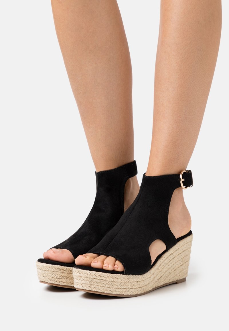 Sandales compensées noires à bout ouvert avec bride cheville et plateforme en jute tressée, portées pieds nus sur un fond uni.