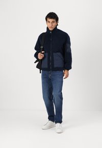 Marineblaue Fleecejacke mit Reißverschluss vorne, hohem Kragen und kontrastierenden dunkelblauen Einsätzen. Quadratische Taschen und ein glänzendes Reißverschlussdetail. Weiße Sneakers getragen.