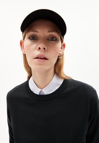 Schwarzer Strickpullover mit rundem Halsausschnitt, getragen über einem weißen Hemd mit Kragen. Eine schwarze Mütze sitzt darauf, aus glattem Stoff und mit einem gebogenen Schirm.