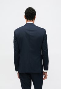 Sötét tengerészkék, szabott öltönyzakó éles hajtókákkal, hátul egyetlen sliccel és strukturált szabással, hozzá illő nadrággal párosítva.