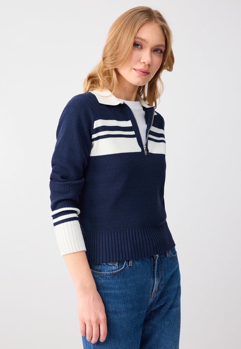 Femme aux cheveux blonds portant un pull zippé à rayures bleu marine et blanc ainsi qu'un jean bleu, sur un fond uni gris clair.