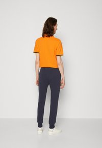Oranges kurzärmliges T-Shirt mit marineblauen Akzenten, kombiniert mit marineblauen tapered Pants und weißen Schuhen, von hinten vor einem grauen Hintergrund betrachtet.