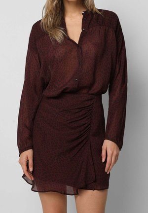 Femme portant une robe à manches longues, rouge foncé à imprimé léopard, avec un détail froncé sur la jupe et un col en V boutonnié.