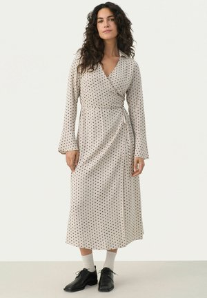 Beige-kietova mekko mustilla pilkuilla, pitkät hihat ja kaulus, jossa on vyötäröllä solmittava sulkija ja keskipitkä leikkaus.