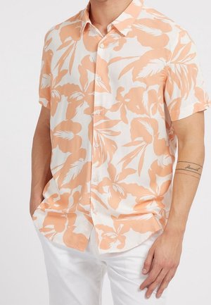 Homme portant une chemise à manches courtes boutonnée avec imprimé floral pêche et un pantalon blanc, debout les mains dans les poches sur fond blanc.