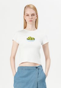 Balta trumpa rankovėmis crop top tipo palaidinė, kuriame priekyje pavaizduotos žalios alyvuogės dubenyje, derinama su mėlynais džinsiniais šortais.