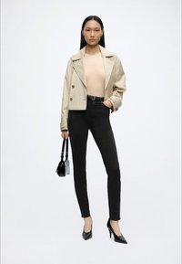 Kvinde i beige cropped jakke, beige top, sorte højtaljede skinny bukser, sorte hæle, der holder en lille sort håndtaske mod hvid baggrund.