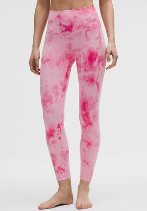 Osoba ubrana w wysokie legginsy w różowo-czerwony wzór tie-dye, stojąca boso na jednolitym tle.