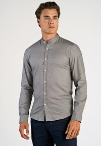 Uomo che indossa una camicia grigia a colletto alla mandarina aderente con bottoni bianchi e pantaloni scuri, mentre guarda di lato su uno sfondo semplice.