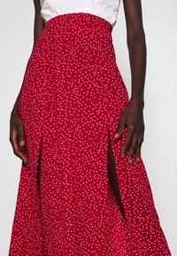 Jupe rouge à pois blancs, à taille haute, avec deux fentes latérales et un tissu fluide qui tombe jusqu'au sol. Design texturé.