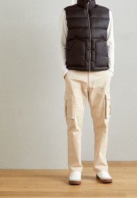 Person som bär en svart pufferväst, vit långärmad tröja, beige cargobyxor med sidofickor och vita sneakers på ett trägolv.