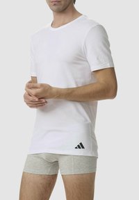 T-shirt bianca in cotone con colletto rotondo e maniche corte, con un logo Adidas nero nella parte inferiore sinistra. Dei pantaloni boxer grigi sono visibili sotto.