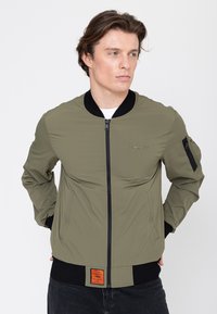 Junger Mann trägt eine olivgrüne Bomberjacke mit schwarzem Kragen und Bündchen, Hände hinter dem Rücken, blickt nach rechts.