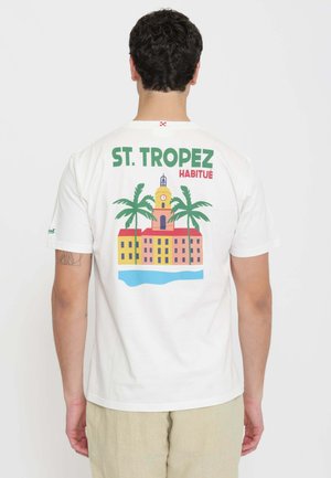 Uomo che indossa una maglietta bianca con disegno colorato di edificio di Saint-Tropez e palme sulla schiena, pantaloni beige, in piedi di spalle.