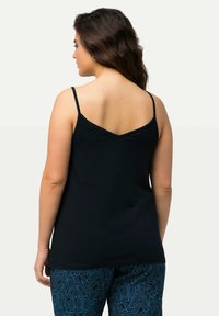 Černý top bez rukávů s tenkými ramínky, se vzadu ve tvaru písmene V. Kombinován s modrými kalhotami s potiskem, zobrazujícími paisley vzor.