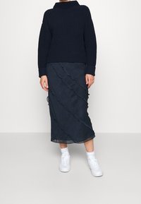 Granatowy prążkowany sweter z kołnierzem typu komin, zestawiony z dopasowaną falbanianą spódnicą z lekkiego materiału oraz białymi trampkami i skarpetkami sięgającymi kostek.