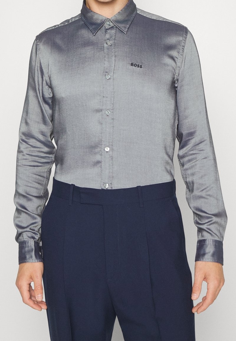 Homme portant une chemise grise brillante à boutons avec le logo "BOSS" sur la poitrine et un pantalon taille haute bleu foncé, debout devant un fond uni.