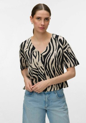 Kobieta o ciemnych włosach związanych do tyłu, ubrana w czarno-beżową koszulę na krótki rękaw w paski zebry oraz jasnoniebieskie dżinsy.