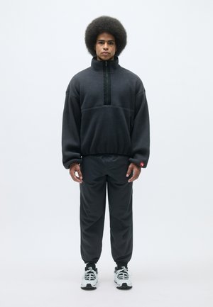 Sötétszürke fleece pulóver, félcipzáras kialakítással, fekete cipzárral és zsebbel. Fekete tapered nadrággal és szürke sportcipővel párosítva.