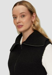 Schwarze Strickweste mit hohem Kragen und Reißverschluss, getragen über einem weißen Langarmshirt, mit geripptem Muster.