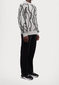 Sudadera gris con patrón de cadena negra y texto, con puños y dobladillo acanalados, combinada con pantalones negros y zapatillas con acentos blancos.