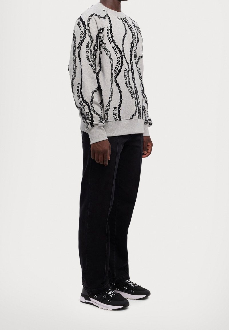 Sudadera gris con patrón de cadena negra y texto, con puños y dobladillo acanalados, combinada con pantalones negros y zapatillas con acentos blancos.