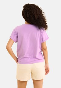 T-shirt lilas à manches courtes en coton doux, présentant une coupe décontractée et des ourlets roulés, assortie à un short beige clair avec des poches arrière.