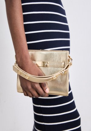 Donkere hand die een gouden metallic clutchtas met band vasthoudt, tegen een marineblauwe jurk met witte horizontale strepen.