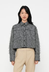 Jonge vrouw met steil zwart haar draagt een grijze, gekleurde cropped blouse met knopen en beige hoge taille broeken tegen een witte achtergrond.