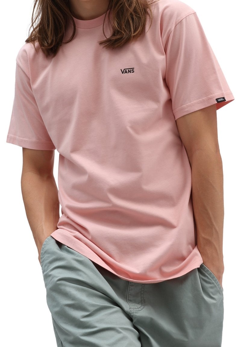 Personne portant un t-shirt Vans rose clair avec le logo sur la poitrine et la manche, associé à un pantalon gris clair, les mains dans les poches.