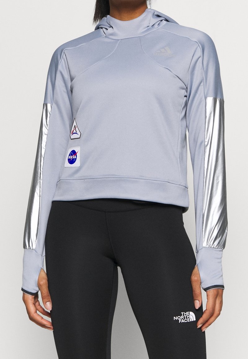 Femme portant un sweat à capuche gris clair avec des écussons NASA et des manches avec des bandes argentées, associé à des leggings noirs avec le logo The North Face.