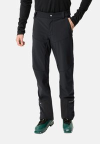 Pantalones de esquí negros con bolsillos y piernas inferiores reforzadas, con una textura suave y un diseño ajustado, combinados con botas de esquí verdes.